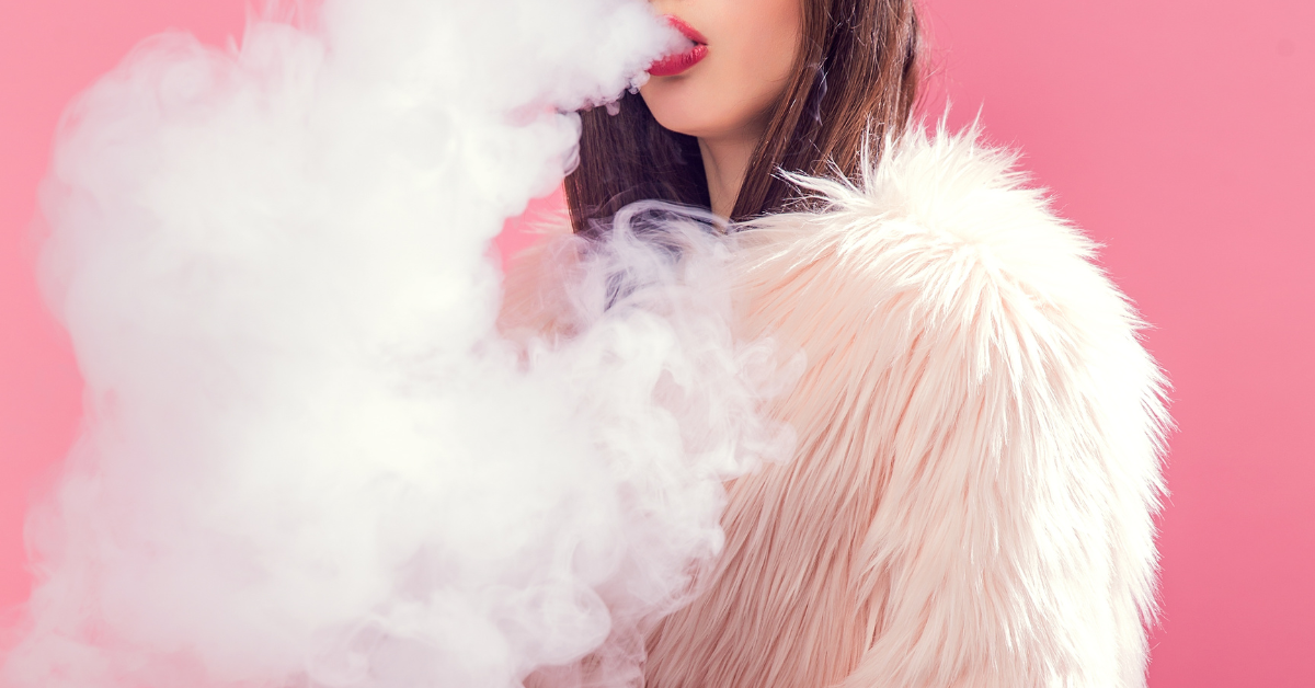 A hot person in an extremely sexy fur coat exhales a cloud of vape smoke against a pink background.